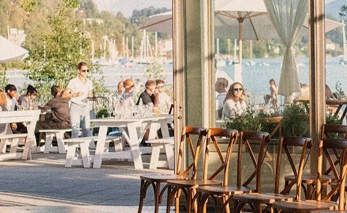 Eine Szene mit Tischen und Stühlen im Freien am See, die unter grossen Sonnenschirmen stehen.