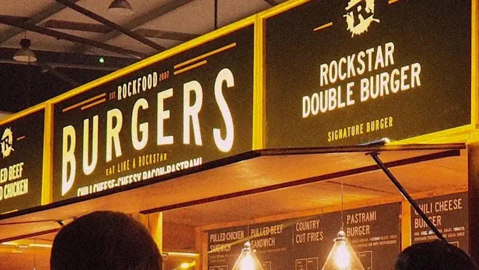 Eine lebhafte Imbissstand-Szene mit einem auffälligen Schild, auf dem ‚Rockfood Burgers‘ und ‚Rockstar Double Burger‘ zu lesen ist. Der Stand strahlt mit seiner warmen Beleuchtung und den rustikalen Holzakzenten eine lebhafte, einladende Atmosphäre aus.